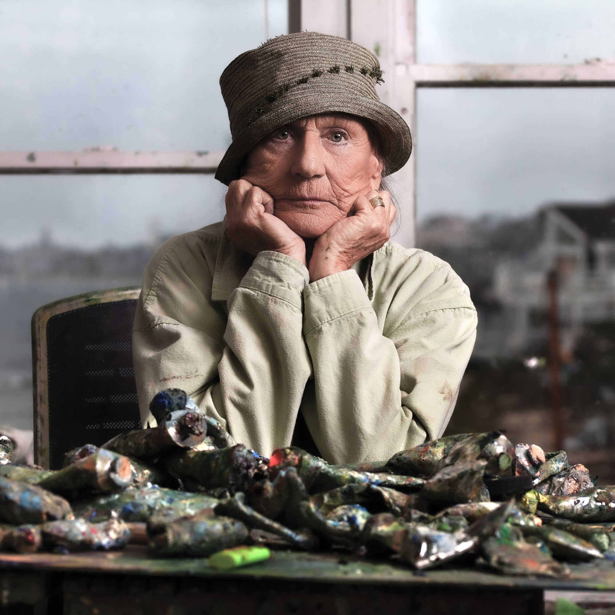 Anne Packard seated behind a table full of old paint tubes in from of windows with a view of Provincetown behind her.