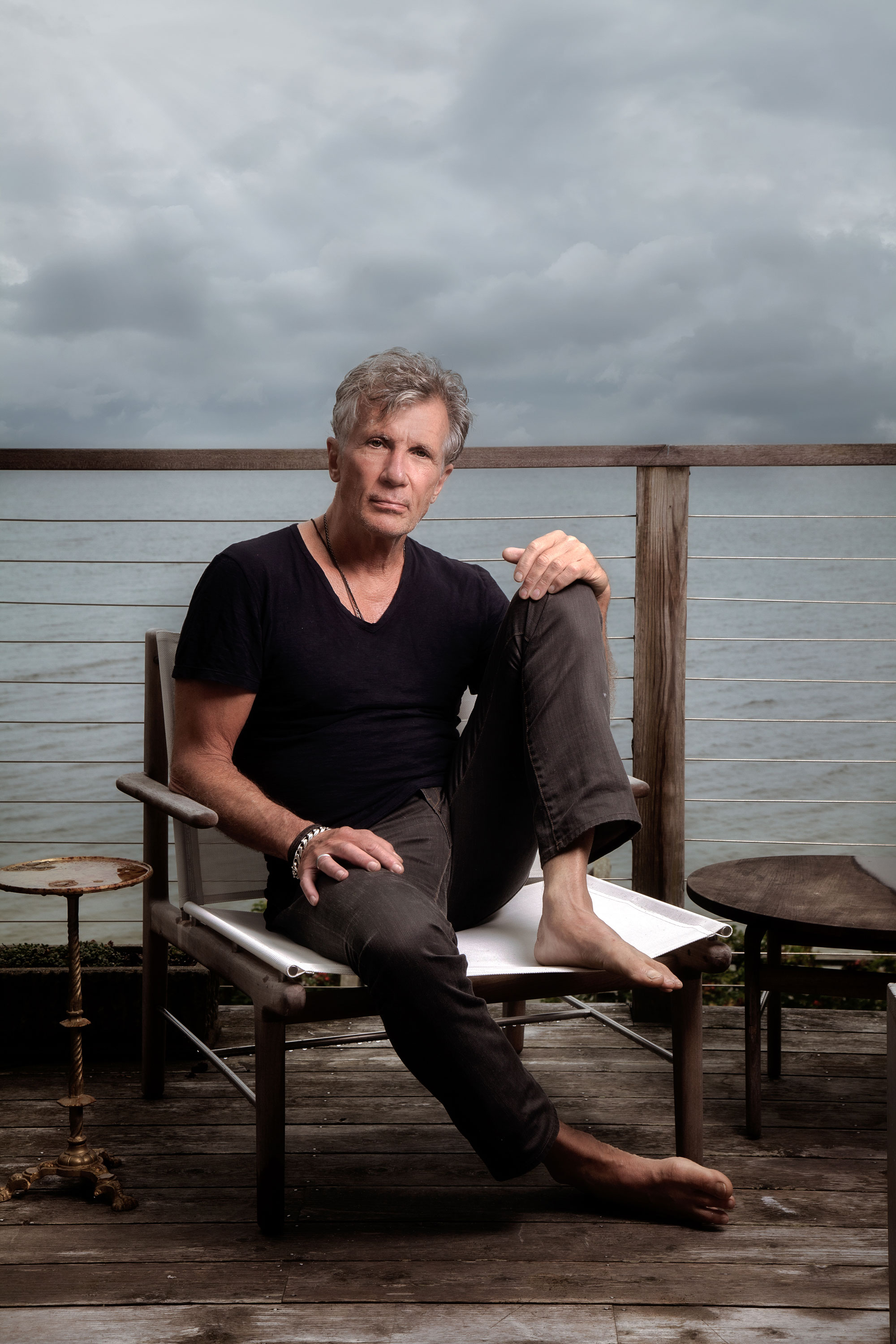 Author Michael Cunningham photographed by Ron Amato on seated on his deck overlooking the ocean, with gray, stormy skies in the background